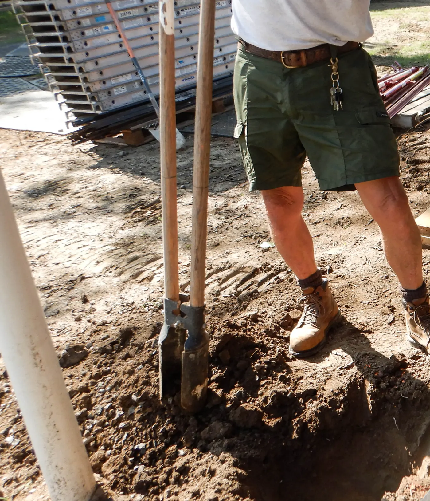 An open post hole dug in bare soil with two long-handled shovels standing in it.