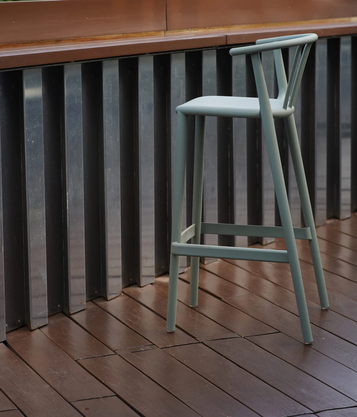 A pale bar stool standing on a dark stained deck beside a slatted screen.